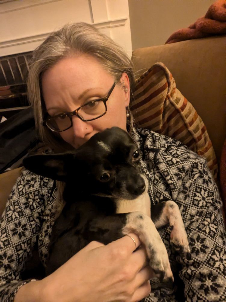 A gray haired woman holds a Chihuahua/ Boston terrier dog. The dog is mostly black, with a white belly and white front paws. The woman's mouth is hidden by the dog's head. Both are snuggling and have expressions of content. 