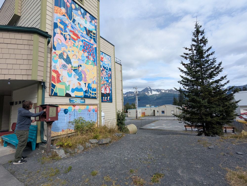 A colorful mural decorates the wall of a building in Seward, AK. Mountains can be seen against a cloudy but blue sky in the background. An evergreen tree is on the right side of the photo. The foreground is an empty parking lot. A person can be seen in the foreground on the left, peering into a little free library kiosk. 