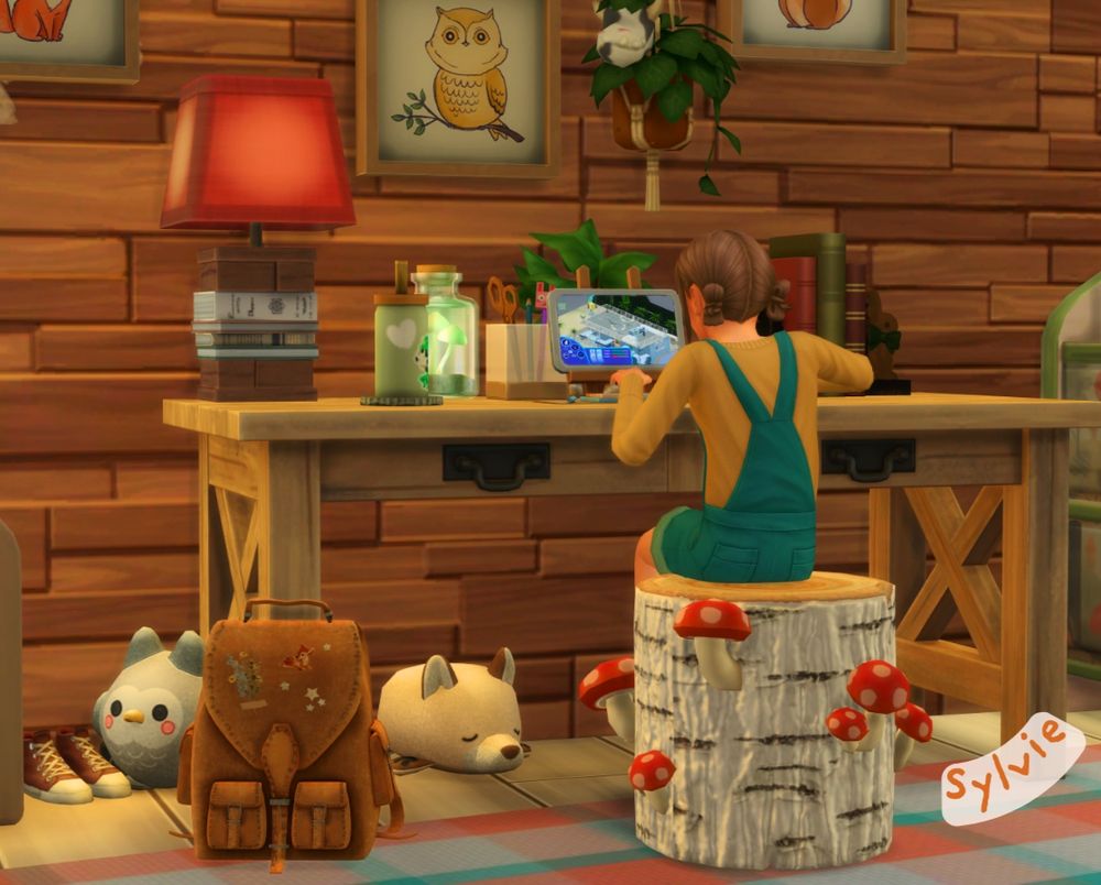 Sylvie sits at her desk, sitting on a white birch bark stool, with red mushrooms sprouting from the sides. On the floor are a couple of stuffed woodland animals, and a brown leather satchel backpack with a couple of nature decals on the flap. On the reclaimed wooden wall are a few framed pictures of woodland animals.