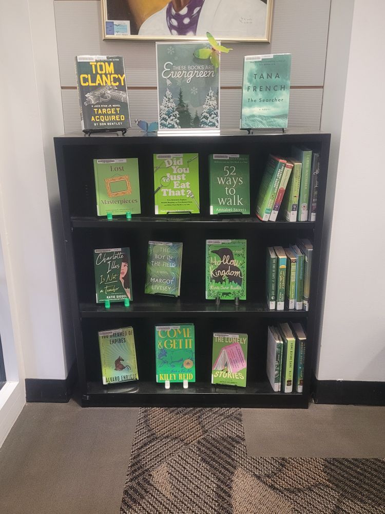 A black three-shelf bookshelf in an alcove with three books facing out and 3-5 books spines-out on each shelf, along with two books on the top of the bookshelf. Also on the top of the bookshelf is a sign that says, "These books are evergreen." And indeed, each book cover and spine is predominantly some shade of green. (There's also a green butterfly attached to the sign; it's left over from the summer reading decorations; we still have most of the butterflies up because they're pretty.)