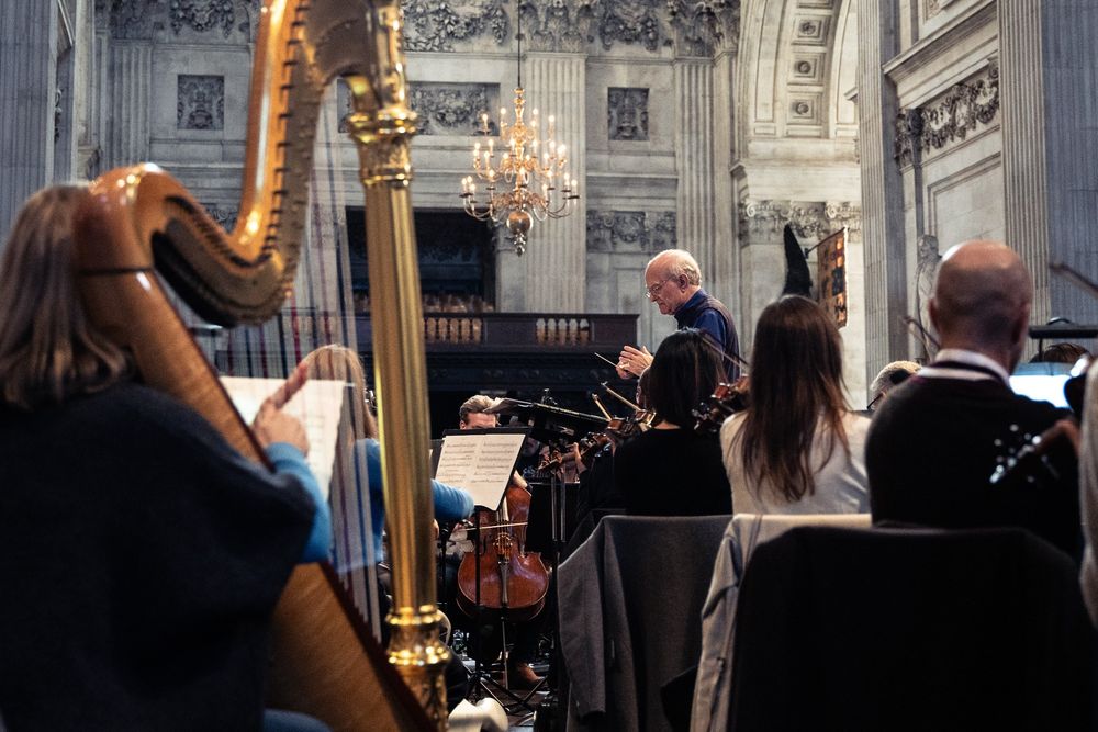 John Rutter conducting the Orchestra in rehearsal in a church, with a harp, cellos and violins visible.