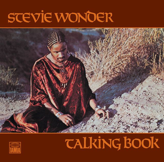 Cover of album "Talking Book" by Stevie Wonder. The top and bottom bars say "Stevie Wonder" and "Talking Book". The latter has a TAMLA logo on the left side. The middle of the cover shows a picture of Stevie on long dark clothes, tied up hair, with his hands on the ground.