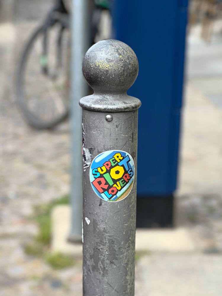 A gray street post with a colorful sticker on the Mario style, which reads Super Riot Lovers. 