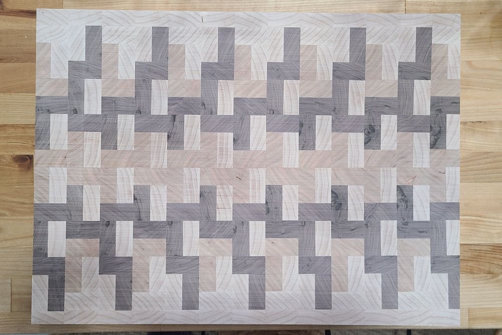 Top view of a walnut, maple, and cherry cutting board sitting on top of a counter. The pattern of the woods is random and geometric.