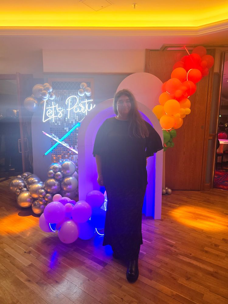 An image of Me in front of various coloured balloons and a neon sign saying "let's party"