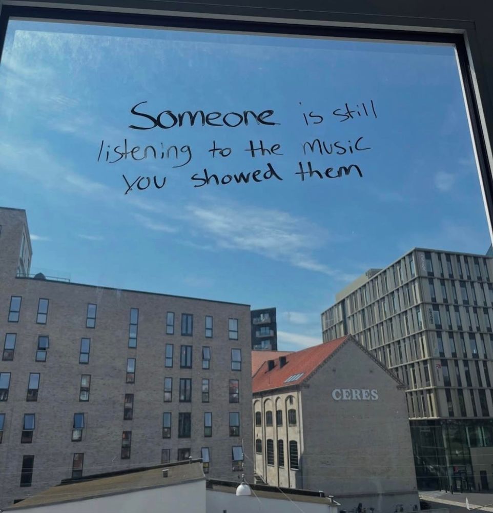 Black sharpie writing on a window, with big tan buildings in the background. The writing, set against the backdrop of a blue sky with wispy clouds, reads: "Someone is still listening to the music you showed them."