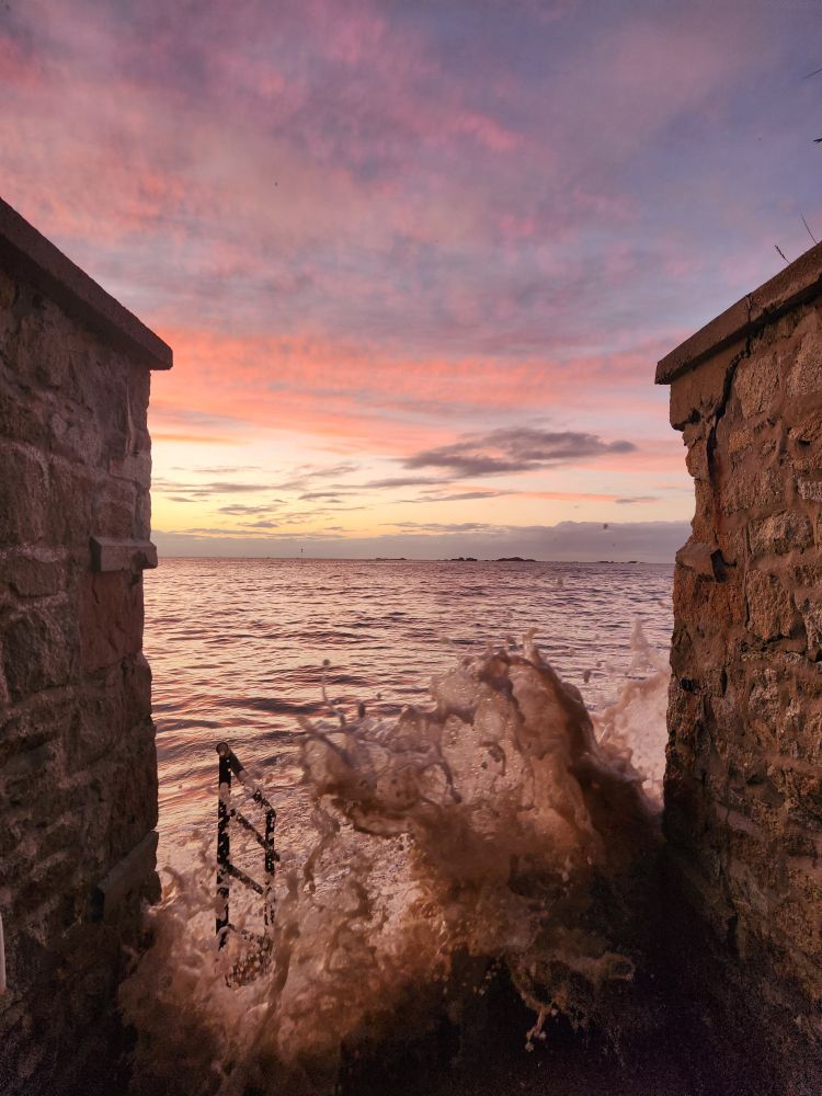 Les vagues s'élancent vers un escalier qui mène à une plage. Le ciel est coloré de jaune, orange, bleu parsemé le soleil couchant. 