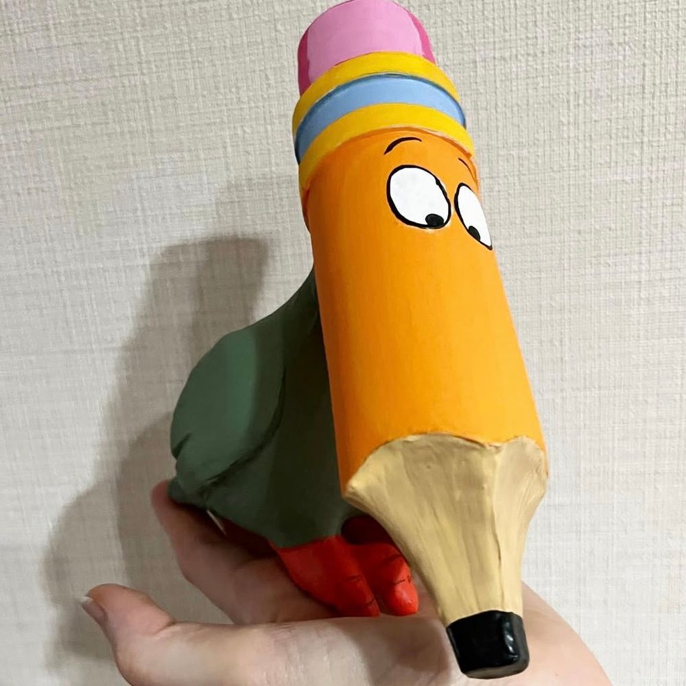 A painted statue of bird like creature with a pencil for a head sitting on a palm against light wall paper