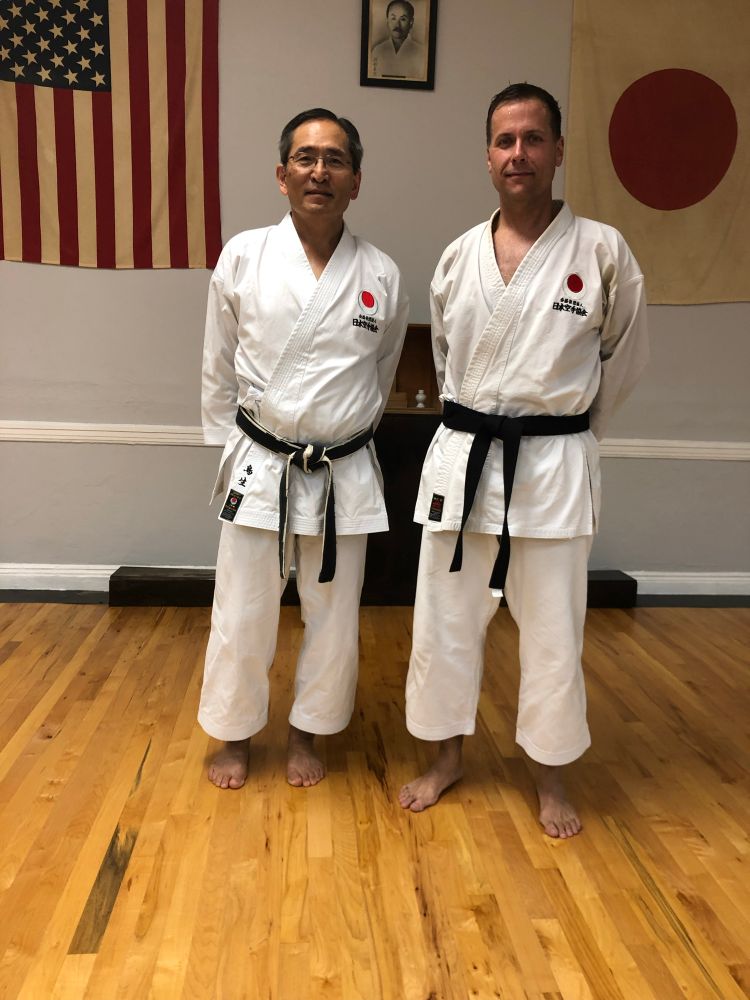 Two black belt karatékas pose for camera in gi at the dojo. Behind, an American and a Japanese flag are on the wall at equal height. 