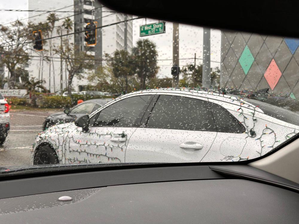 A white sedan with string christmas lights wraped in back and forth patterns over its panels.