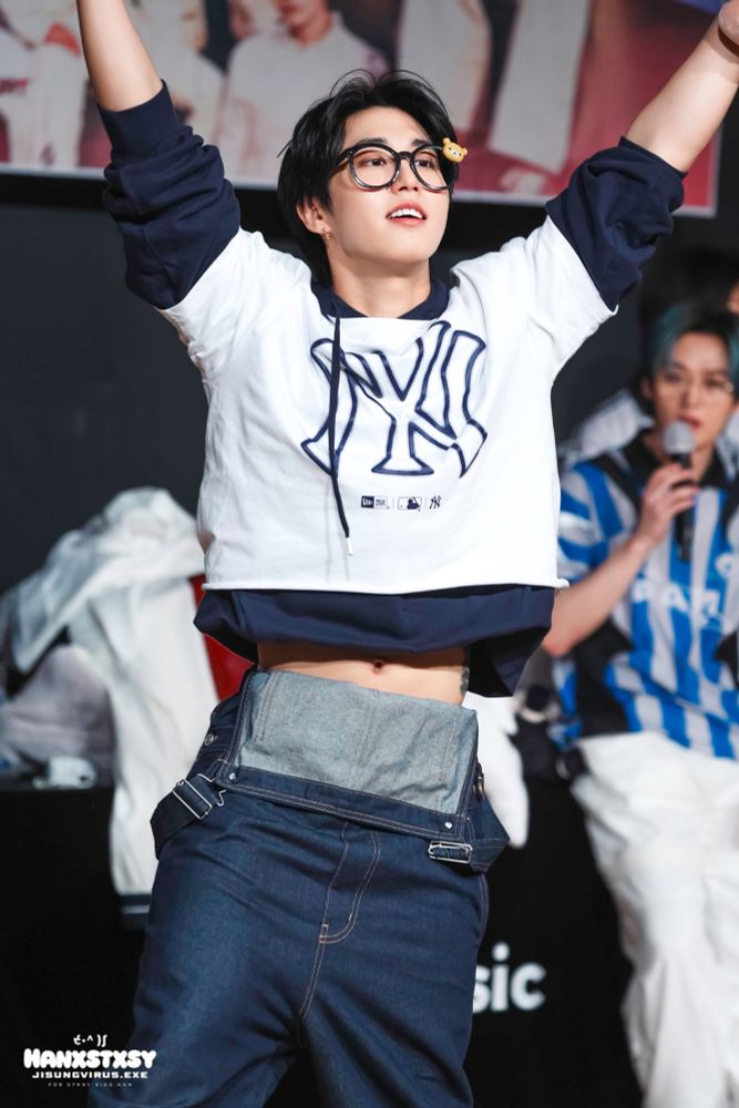 han jisung of stray kids with his hands in the air. he’s wearing thick black rim glasses and a white t with a navy long sleeve underneath and overalls with the straps down.