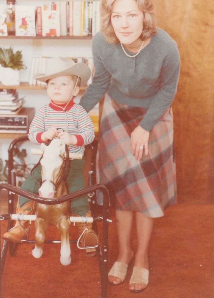 Faded orangey photo of me as a toddler sitting in one of those bouncy horse things with a cowboy hat on. Mom is bending down to stand kinda next to me. She is wearing a plaid skirt and a Donna Reed sweater. This photo was probably taken in 1980 or 1981.