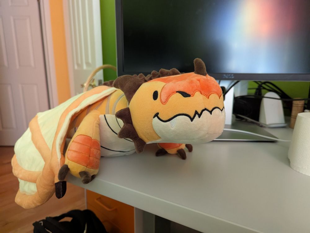 Side profile of a large plushie yellow dragon with orange and brown accents on a desk. He has a lovely smile! 
