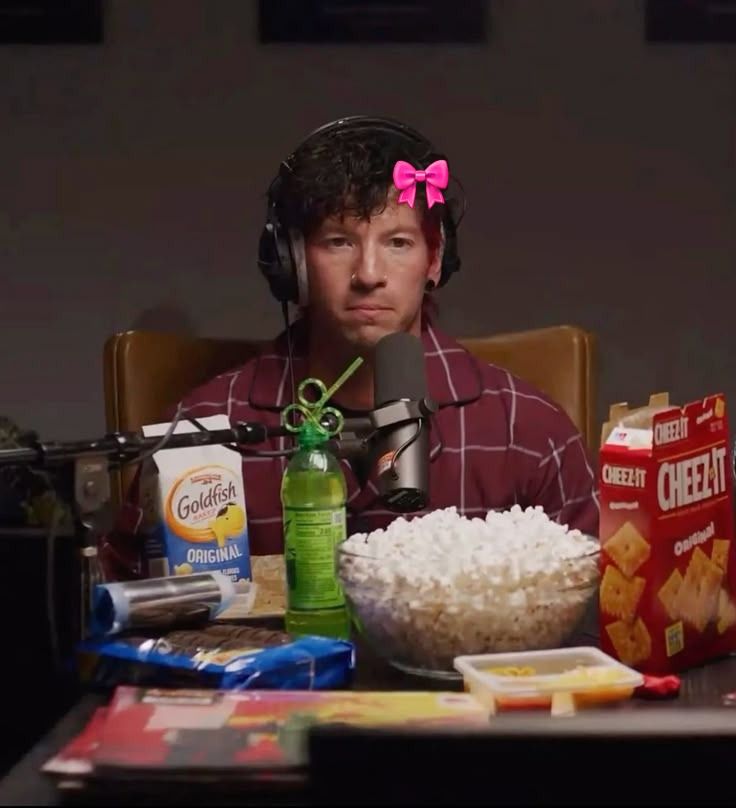 A photo of Josh dun in his pajamas with a bunch of snacks surrounding him. He has a pink bow on his head.