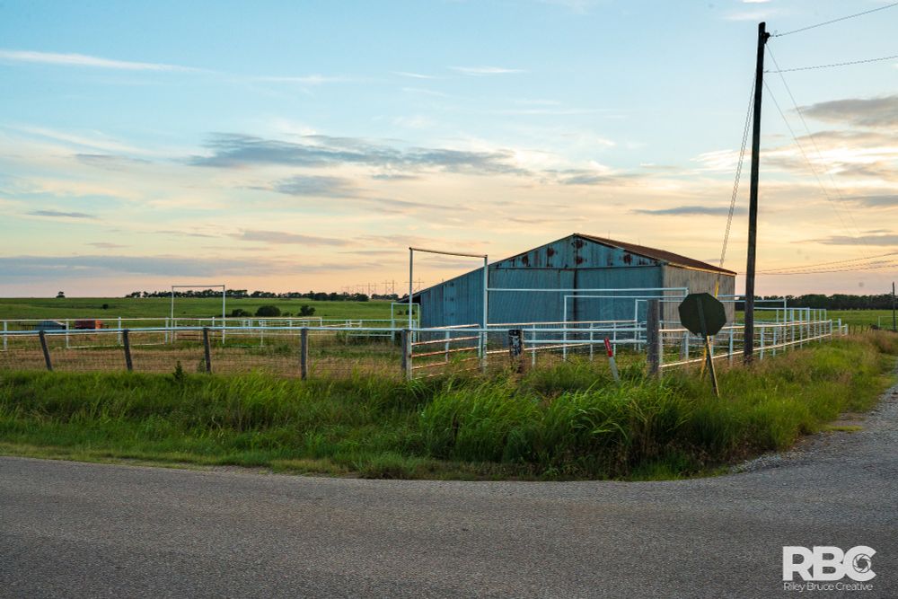 A hay barn at the intersection of a ranch road and a county road in rural Kansas. The sun is setting in the background and there are whips of clouds on the horizon.