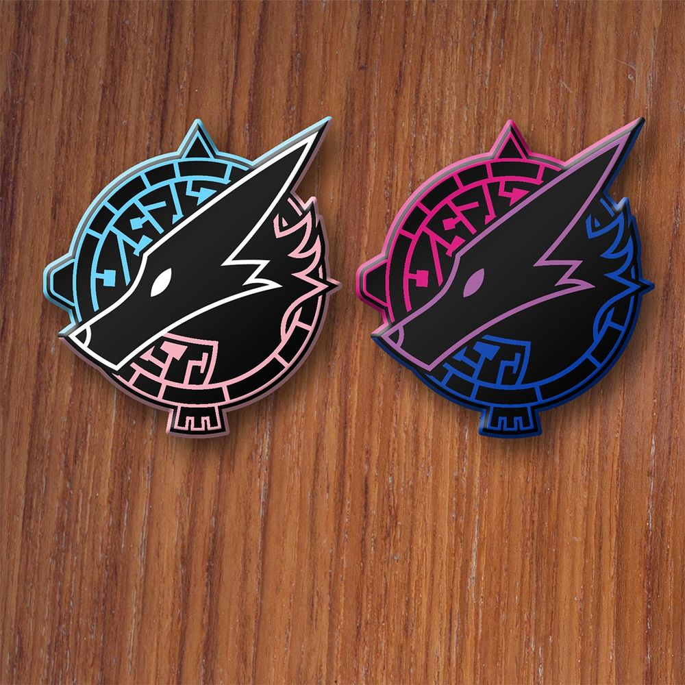 Two mockups of enamel pins featuring the Beast World logo, the head of an undefined beast in a circular field of maze patterns. The pins are in the colors of the trans and bisexual pride flags.
