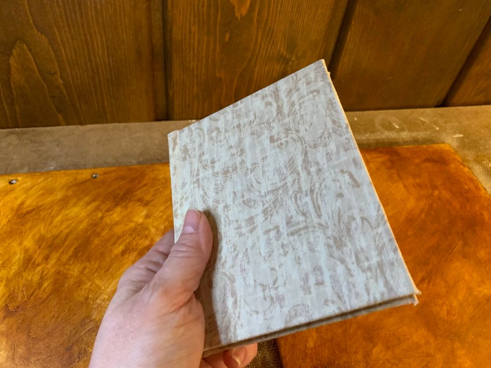 Holding a hand-sized case-bound book. The cover is wrapped in a ticked tan cloth.