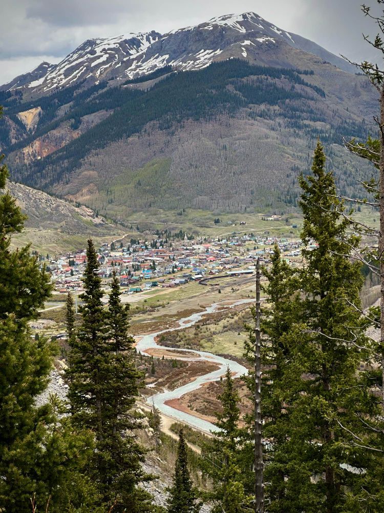 The mountain town of Silverton, CO is seen from a high roadside vantage point, the Animas River snaking down the frame in between nearby pine trees. A bulging foothill looms over the town, its rocky peak patched with snow extending up towards the overcast sky.