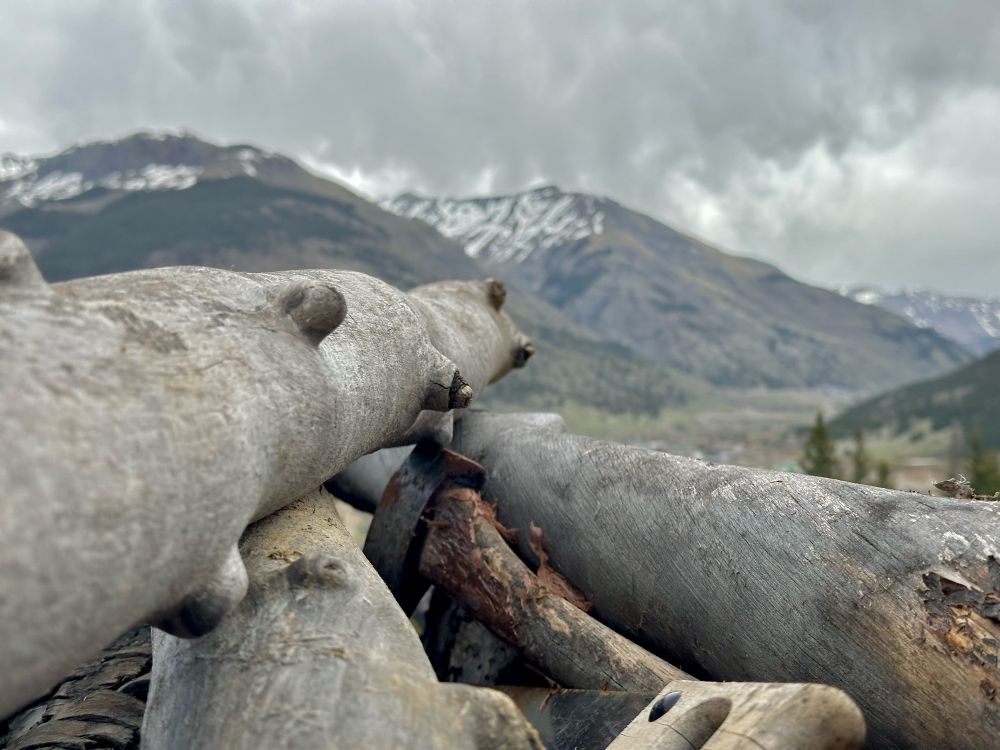 A closely-framed slightly desaturated shot on a pile small logs which are seen along their lengths with an axe and a saw tucked down in between them, both of their handles well weathered. The shallow depth of focus on the woodgrain has the distant low mountains slightly blurred. The sky is all low, stormy clouds that appear to be ready for a light rain shower.