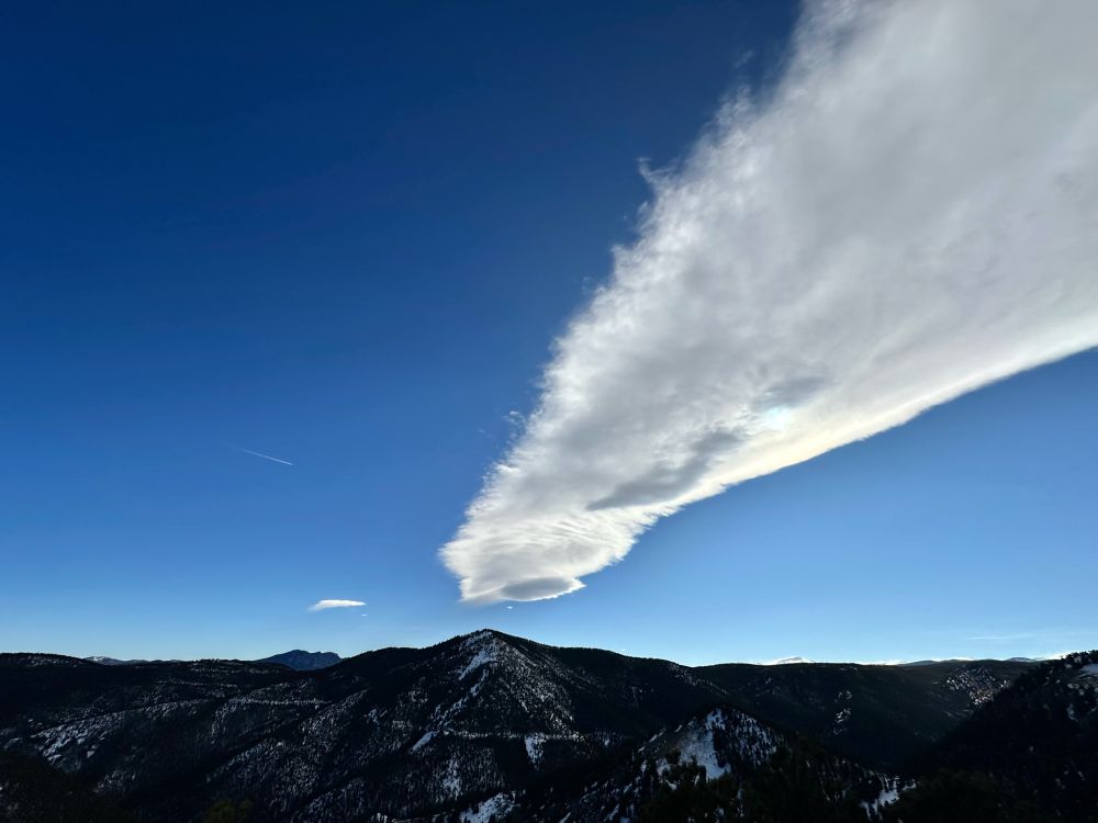 A linear cloud formation that originates above a near-distant foothill peak and continues up and left out of the corner of the frame. The cloud has a smooth right edge and an uneven left edge. The peak only just rises above the continuous line of distant hills, and is centered horizontally while the silhouetted hills are aligned along the lower third of the image. Patches of snow are visible on the hillsides. The sky is otherwise cloudless and has a gradient of blue hues from very light along the hills to a very deep and dark at the top of the photo.