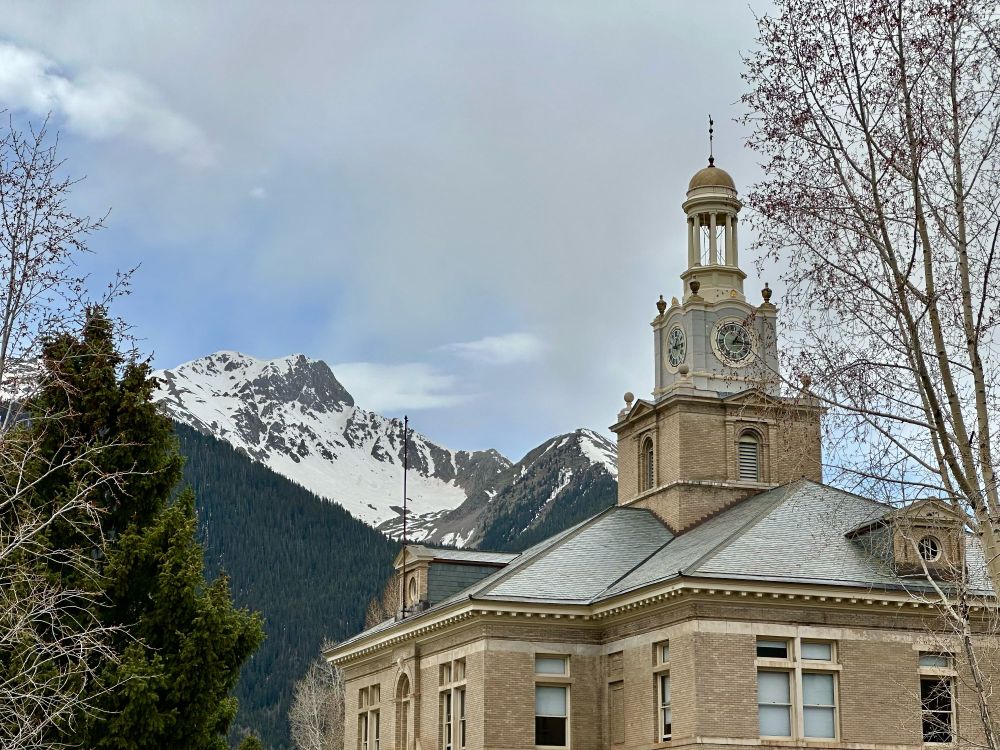 The upper floor windows, roof, and clock tower of the San Juan County Courthouse are seen right hand foreground with the (mostly) snow-covered peak of Sultan Mountain clearly visible behind and to the left. The image is framed by two bony, as-yet-unbloomed aspens and a single pine tree, along with a glimpse of a densely pine treed foothill.