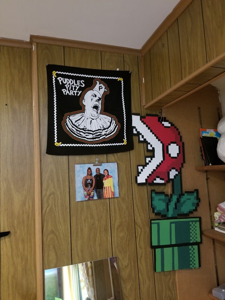 A bandana with a peirot clown with a tragic face tacked to a wood paneled wall. It reads 'Puddles Pity Party.' 