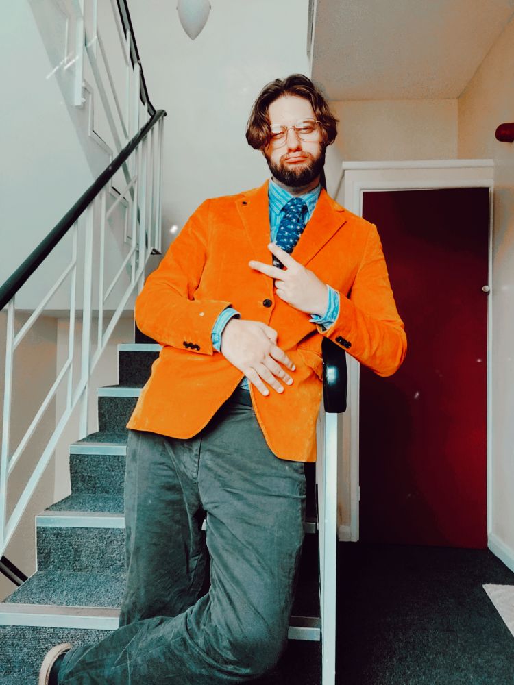 Luke wearing a mustard blazer, blue striped shirt and brown trousers. He has a tie that has small smiley suns and sad overcast clouds on it.