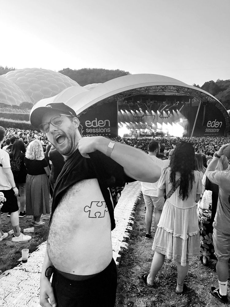 Luke stands in front of the band Biffy Clyro playing music on a large stage at the Eden Project. Luke is holding up his t-shirt to show his Biffy Clyro-inspired puzzle piece tattoo on his ribs.