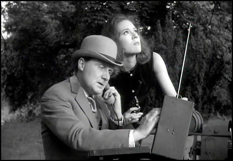 John Steed and Emma Peel using a radio in the Acenters tv show from the 1960s.  Black and white photo.  Steed played by Patrick Macnee in a bowler hat and suit is dialing in the radio and has a phone receiver up to his ear. Peel played by Diana Rigg leans over Steed while looking to the sky. 