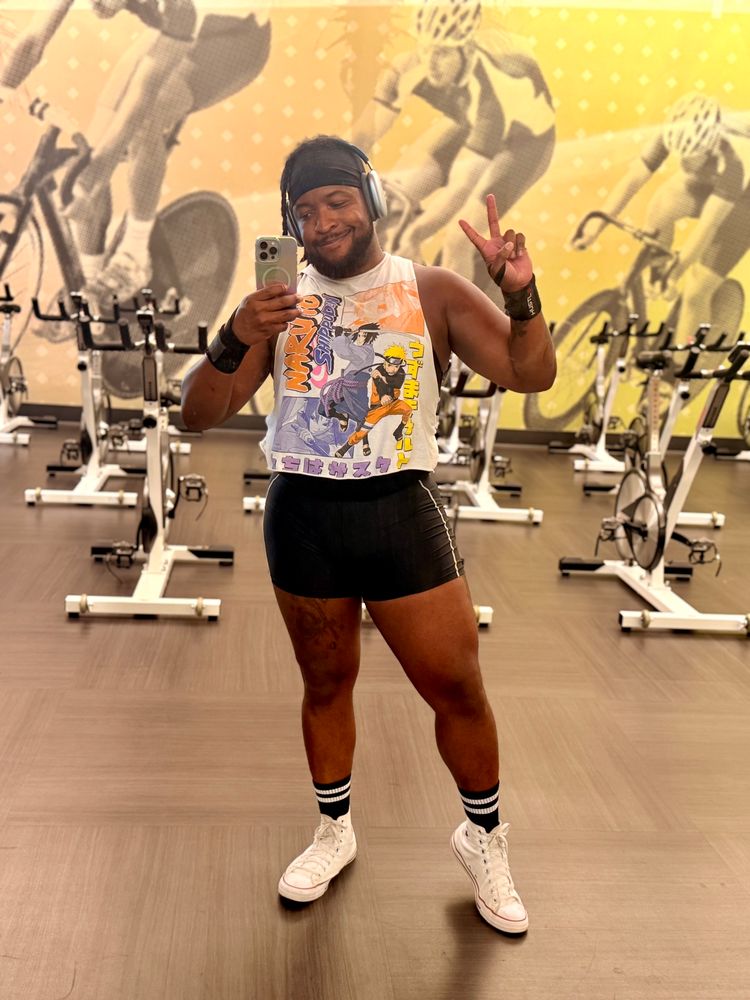Picture of me posing in the gym holding up a peace sign wearing a white Naruto crop top, black shorts, black socks and white shoes.