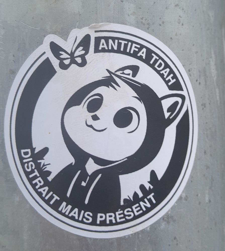 Sticker rond noir et blanc représentant un chat anthropomorphisé dans un style manga, portant un sweat à capuche noir et regardant un papillon. 
Autour, le texte
 "ANTIFA TDAH 
 DISTRAIT MAIS PRÉSENT "