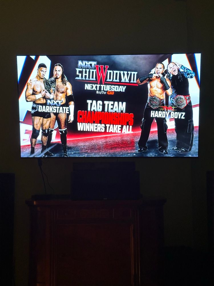 A picture of a television in a dim room showing professional wrestling show WWE NXT. It depicts a tag team championship match where the winners take all between NXT’s DarkState and TNA’s Hardy Boyz.