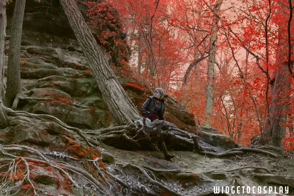 widgetoc, dressed as Nightsister Merrin from Jedi Survivor, is sitting in the roots of an old tree that leans heavily to the left, the roots pulled up and exposed. She is alone in the shot, small in the landscape. The trees have red leaves against a muted orangey-red sky, and the landscape around the tree barren stone and more tangled roots, simulating the planet Dathomir, the home of the Nightsisters.