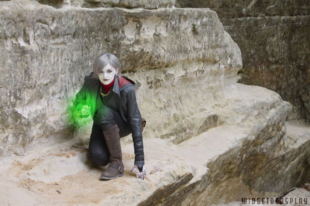 Cosplayer widgetoc as Nightsister Merrin from Jedi: Survivor. She crouches on a rocky ledge in a desert environment. Her right hand is lifted, summoning a swirl of green Nightsister Magick. 