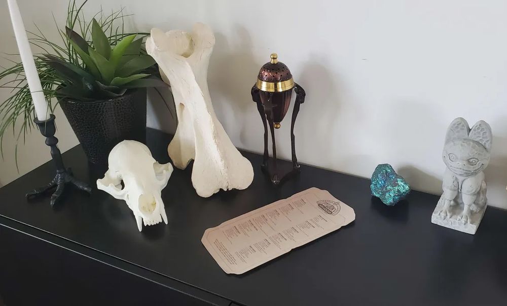 One corner of a long sideboard that features an ecclectic bunch of Jedi Survivor / Pyloon's Saloon-inspired decor, including: a bird-foot candlestick, a resin printed bear skull, a real giraffe vertebrae, a funky little incense burner on long spindly legs, a Pyloon's Saloon cocktail menu (deisgned and selected by yours truly), some "priorite" (chalcopyrite), and a loth cat tiki mug impoted from Batuu.