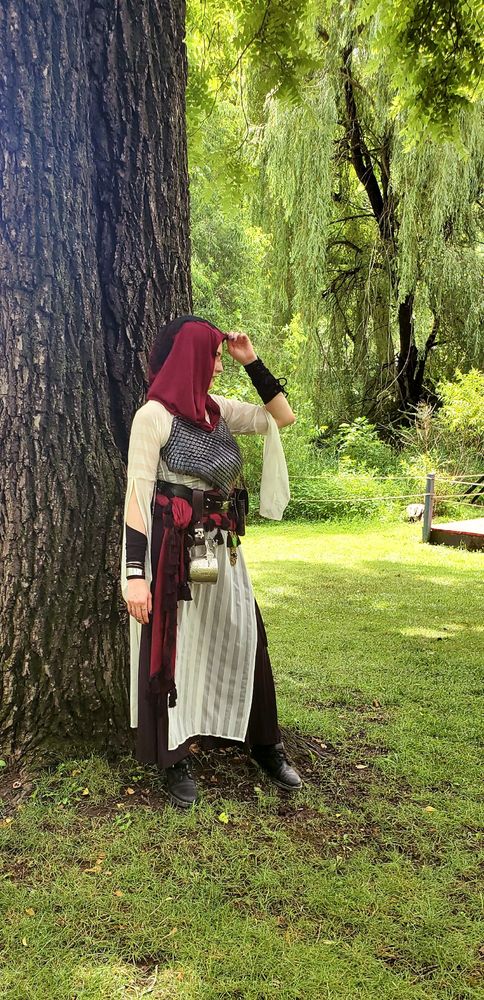 Another shot of widgetoc in her Faire Merrin. She is in profile, showcasing the black lace detail and drape, as well as the sleeves of the white overdress. She has her back to a tree, her left hand raised to adjust the tip of the hood. 