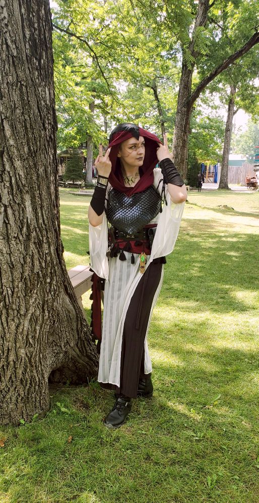 Cosplayer widgetoc dressed as a Renaisaance Faire version of Nightsister Merrin. She has a maroon hood and sash with black detailing, a black scale-mail chest piece reminiscent of Merrin's  leather vest, a white underdress to simulate her skintone, and a loose black pant. She also wears black bracers similar to Merrin's, a silver cuff on her right wrist, and Merrin's iconic necklace and charm. Both hands are lifted to adjust her hood as she glances off to her left, her body slightly crouched and turned to her right. 
