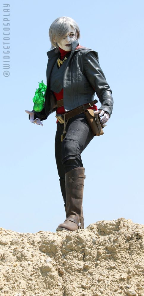 Cosplayer widgetoc dressed as Nightsister Merrin from Jedi Survivor. She is standing atop a rocky hill that looks not unlike the landscape of the planet Jedha. She is looking offscreen to the right with a slight smirk and is conjuring a green flame of Nightsister magick in her right hand. 