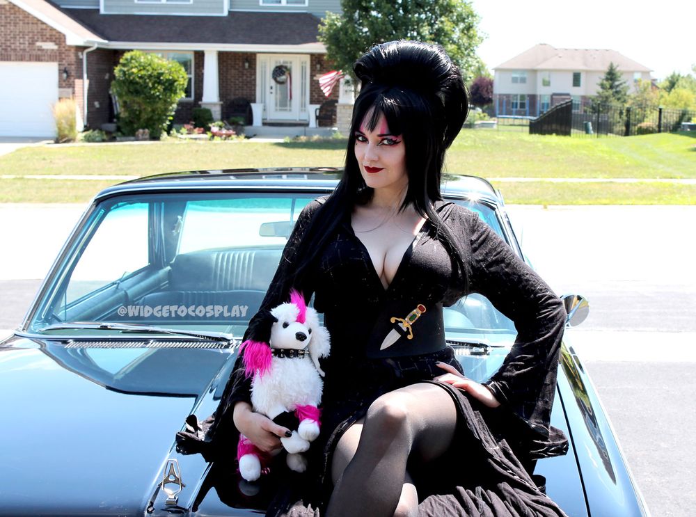 @widgetoc cosplaying as Elvira, complete with big hair, big tits, and big personality. She is sitting on the hood of a black 1965 Dodge to simulate Elvira's Macabre Mobile and is cradling a stuffed version of Elvira's familiar, Algonquin (aka Gonk). 