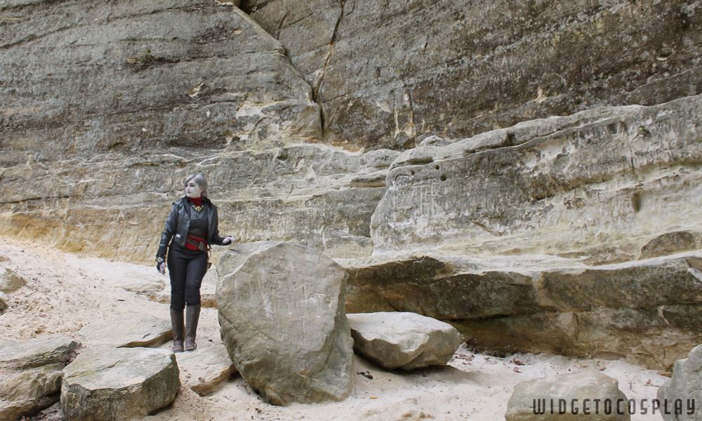Cosplayer widgetoc dressed as Nightsister Merrin from Jedi Survivor. She is in a muted, rocky desert landscape, small in a wide shot that showcases the negative space of the Jedha-like backdrop. She leans on a large rock, glancing offscreen to the left. 