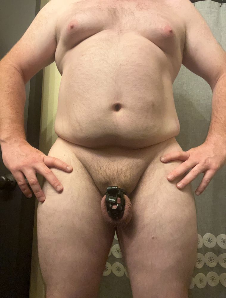 A man standing naked with a black chastity cage around his penis