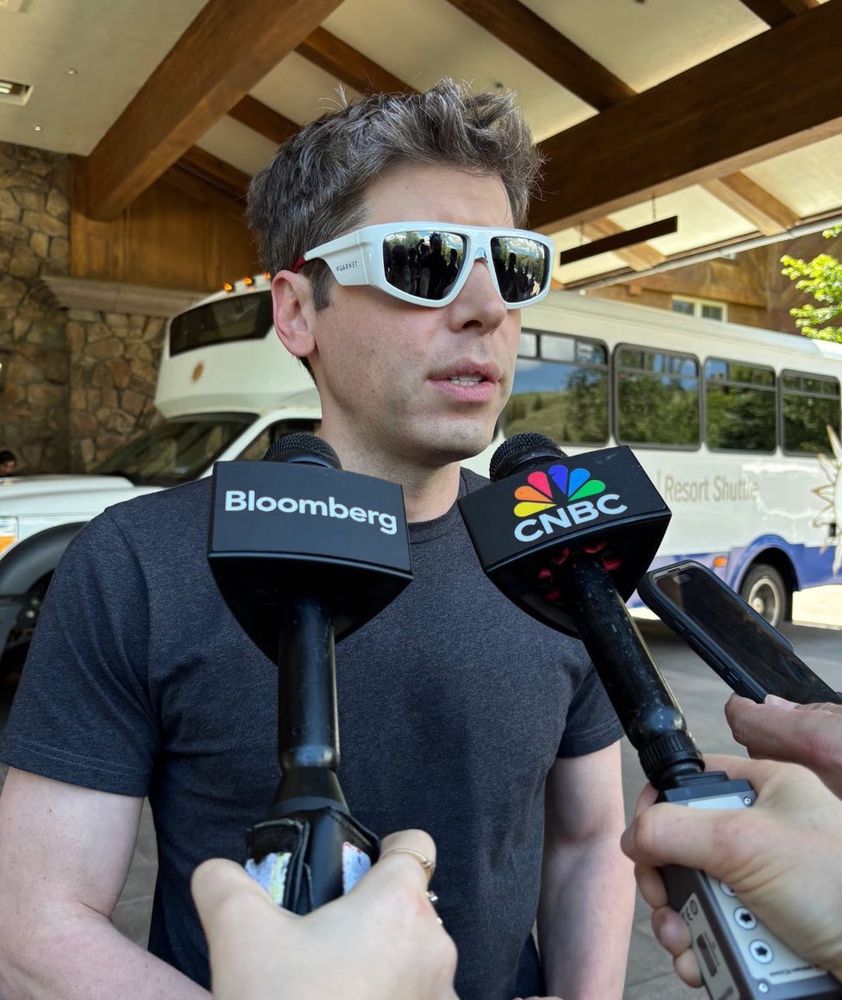 Alt-Text (deutsch):
Ein Mann mit weißer Sonnenbrille wird draußen vor einem Shuttlebus von mehreren Reporter*innen interviewt. Zwei Mikrofone mit den Logos von Bloomberg und CNBC sind auf ihn gerichtet. Er trägt ein dunkelgraues T-Shirt.

Alt-Text (englisch):
A man wearing white sunglasses is being interviewed outdoors in front of a shuttle bus. Two microphones with Bloomberg and CNBC logos are pointed at him. He is wearing a dark grey t-shirt.