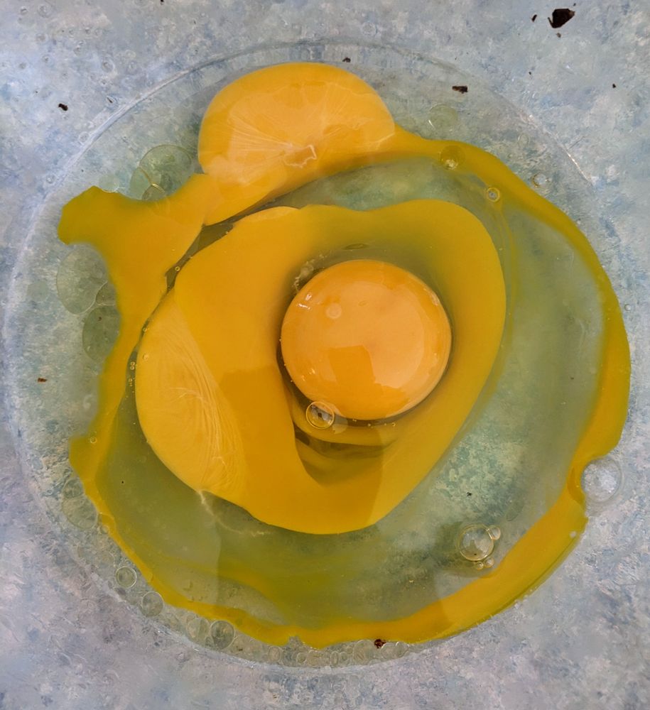 3 Cracked eggs in a glass bowl, seen from above. The yolks form concentric circles. The first yolk broke and the second egg landed right on top of it - pushing the yolk out in all directions. The second yolk also broke and the third egg pushed that yolk out into a circle, but the 3rd yolk did not break - it formed a central yellow ball sorrounded by rings of clear and yellow egg slime