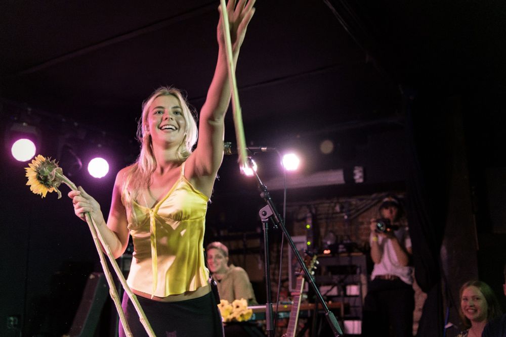 Bebe Stockwell on stage at Mercury Lounge holding plastic sunflowers with long stems in her right hand and throwing one into the audience with her left hand.