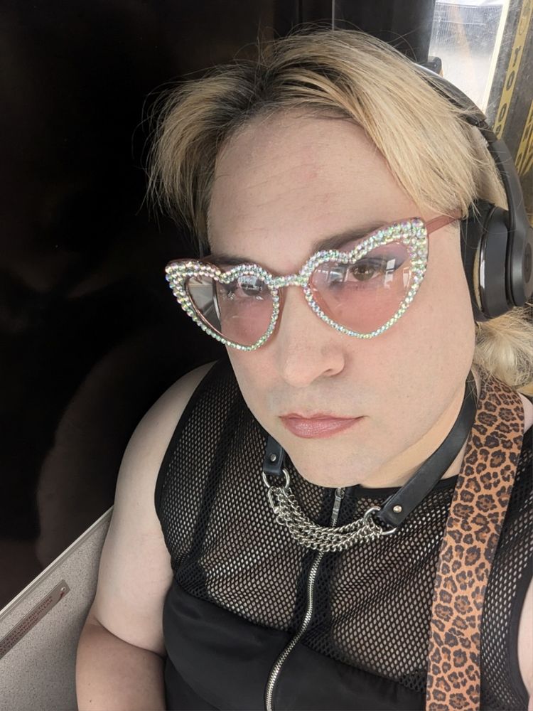 Blonde in black dress looks to the camera. They're wearing a chain and leather collar, headphones, and heart shaped rhinestoned sunglasses.