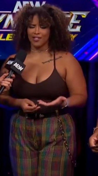 Screen capture of Willow Nightingale, a powerful woman (the babe with the power, if you will), speaks into a microphone. She wears a dark tank top and rainbow plaid pants with a wallet chain.