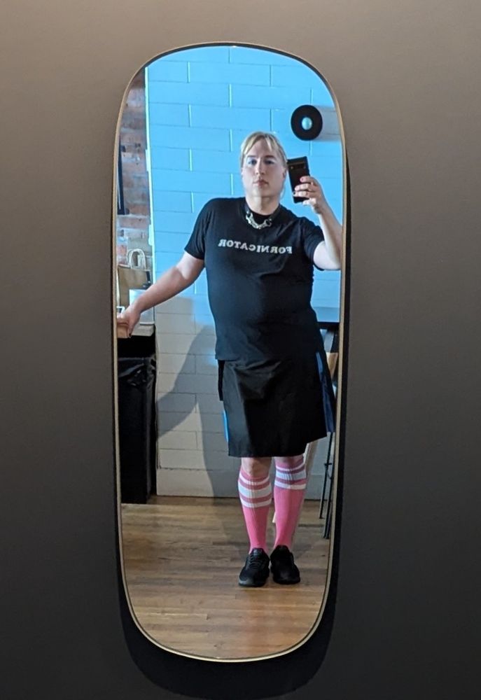 Mirror selfie of a blonde in a black kilt, pink knee high athletic socks, a leather/chain collar and a black T-shirt that reads "Fornicator"