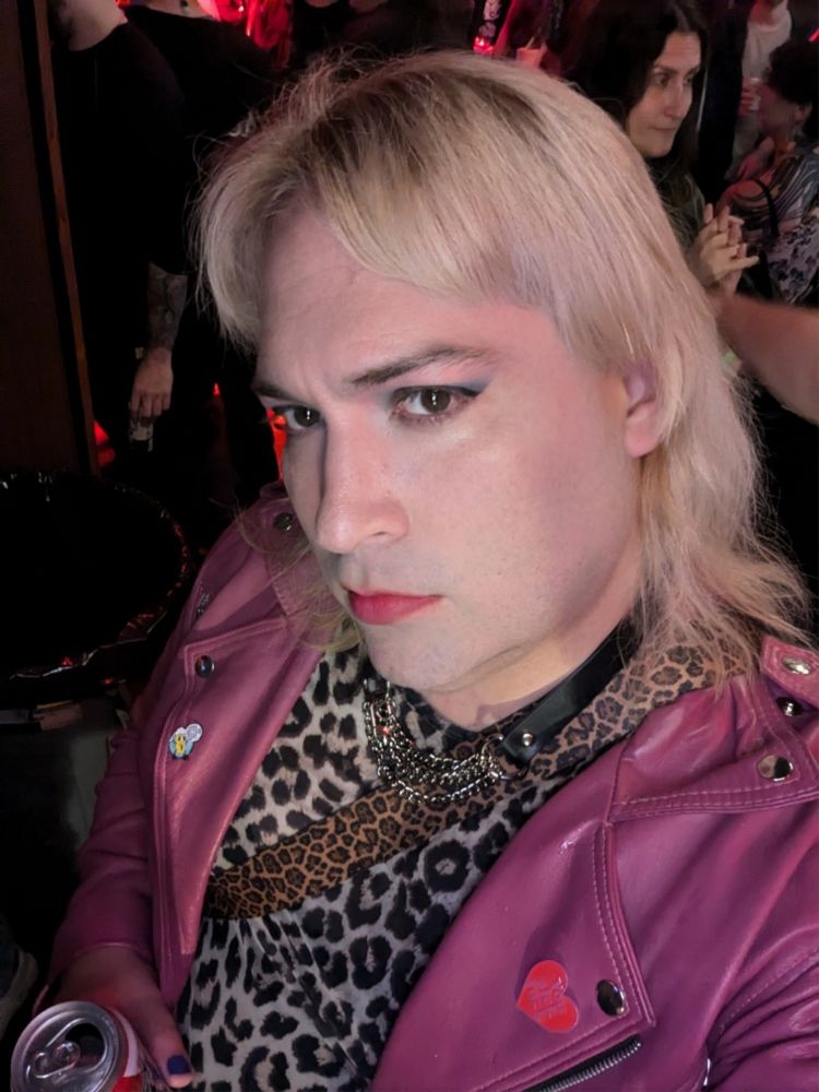 Blonde with smokey eye makeup wearing a lilac colored moto jacket and leather and chain collar, with a leopard print top. They have a beer canning their hand. A crowded music venue in the background.