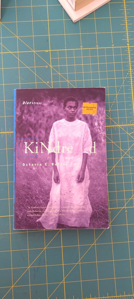Boot cover: Kindred, by Octavia E. Butler. A sepia toned photo of a black woman in a floor length white shift against a background of roughly cut grass, looking fearfully off to the side. 
