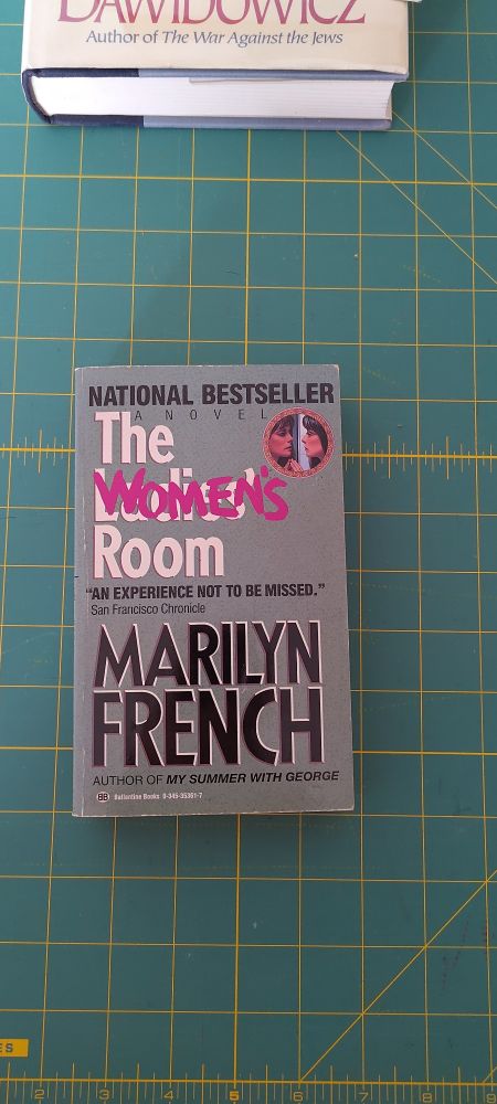 Book cover:The Women's Room, Marilyn French. Grey background. Title in white block font EXCEPT "Women's" in pink handwriting over crossed out "Ladies'". Author's name in black. 